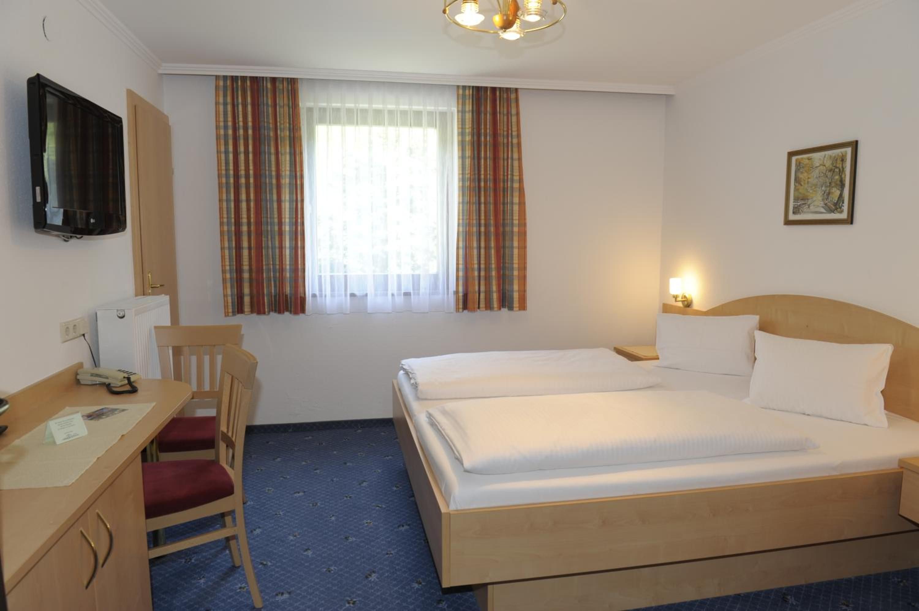 A hotel room with a bed, a desk with a telephone, a chair, a television mounted on the wall, and a window with curtains.