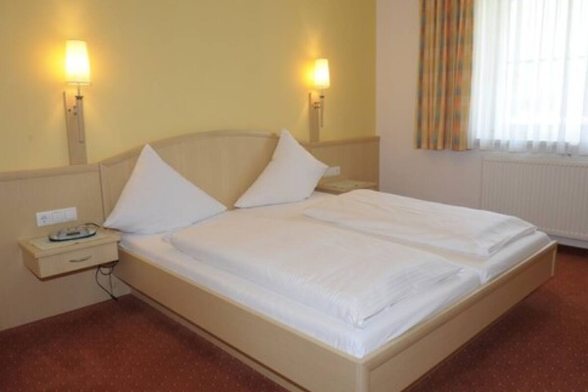 Una cama individual con ropa de cama blanca en una habitación con dos lámparas de noche, una mesita de noche y un ventanal con cortinas.