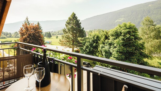 Un balcon avec une table et deux verres à vin, donnant sur un paysage luxuriant, avec des arbres et une montagne lointaine.