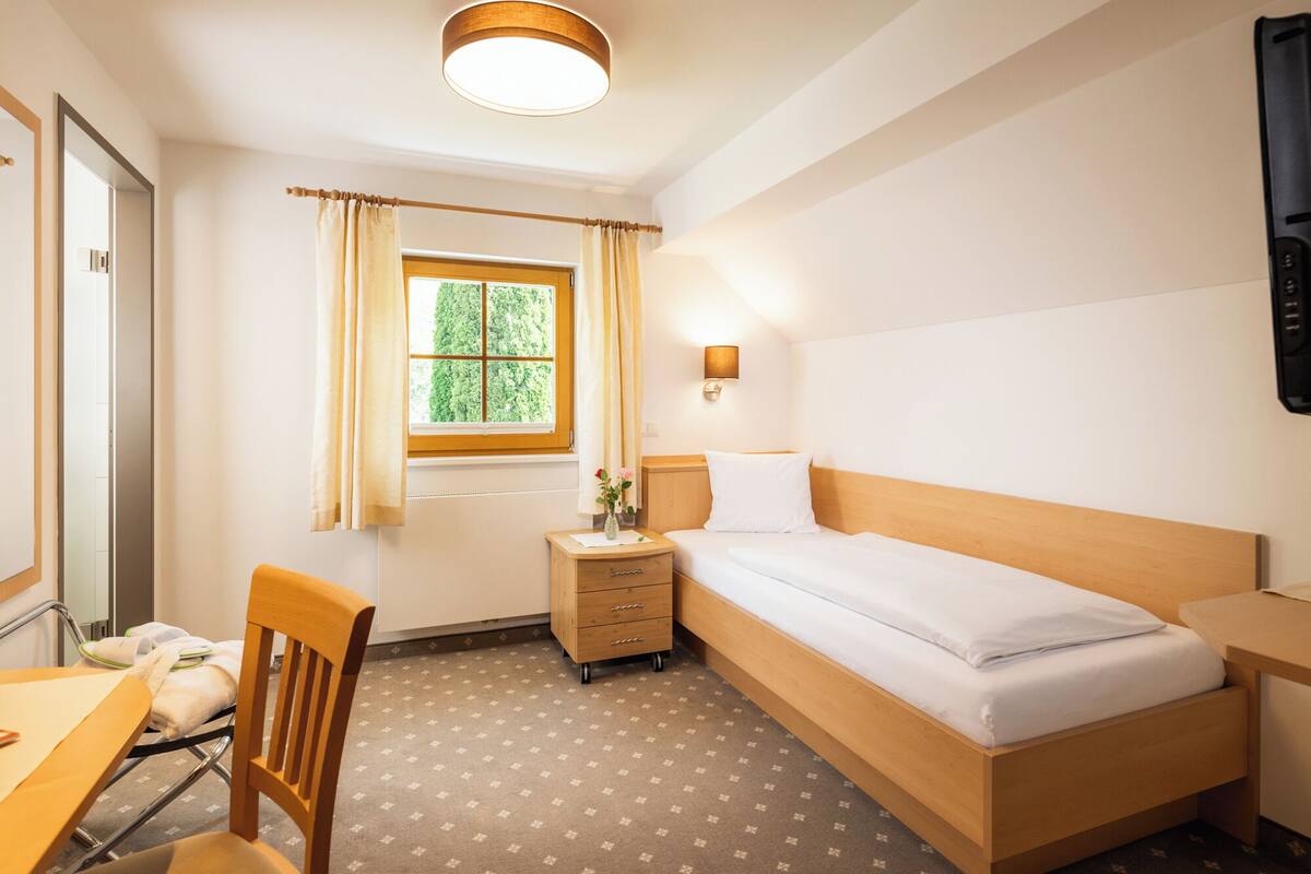 Habitación de hotel con una cama individual, un escritorio, una silla, una mesita de noche, una lámpara y una ventana con cortinas.