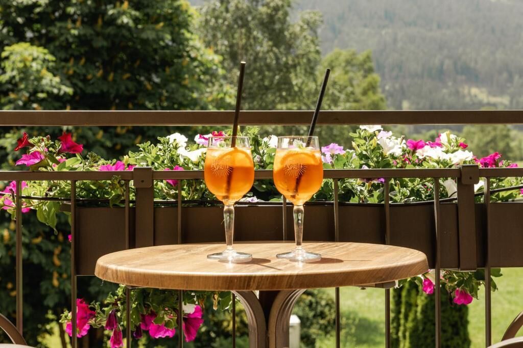 Una mesa de madera redonda con dos vasos de bebida de naranja en un balcón con barandilla metálica y macetas con flores.