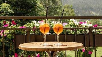 Una mesa de madera redonda con dos vasos de bebida de naranja en un balcón con barandilla metálica y macetas con flores.