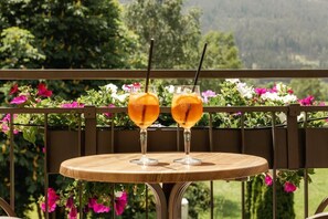 Une table ronde en bois, sur laquelle reposent deux verres contenant une boisson à l’orange, est placée sur un balcon doté d’une rambarde en métal et de jardinières.