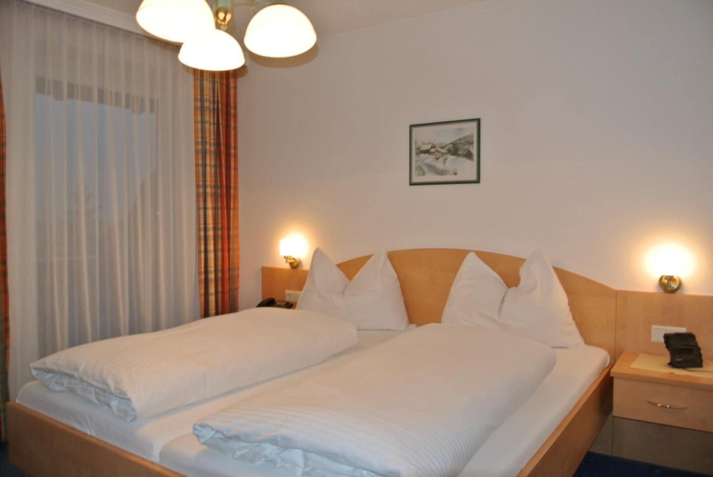 A double bed with white linens, a wooden headboard, two bedside lamps, a nightstand, and a framed picture on the wall.