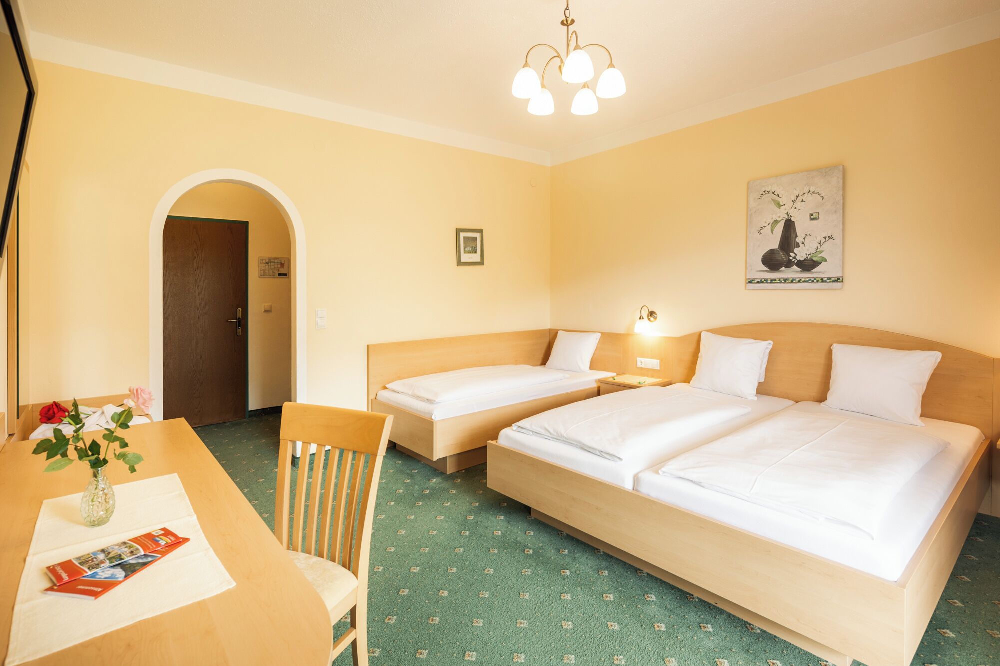 A hotel room with two single beds, a wooden desk with a chair, a small table with a vase of flowers, and a wall-mounted lamp.