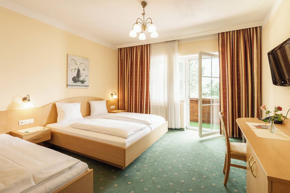 Habitación de hotel con cama, mesitas de noche, escritorio y una silla. Hay una ventana con cortinas, una puerta que da a un área exterior y un televisor instalado en la pared.