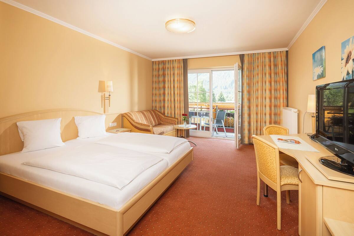 Una habitación de hotel con una cama grande, un escritorio con una silla, un televisor y un balcón con cortinas.