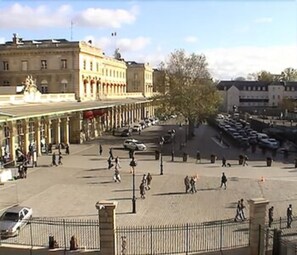 Una amplia plaza pública con una fuente, rodeada de arquitectura clásica y una fila de coches aparcados.