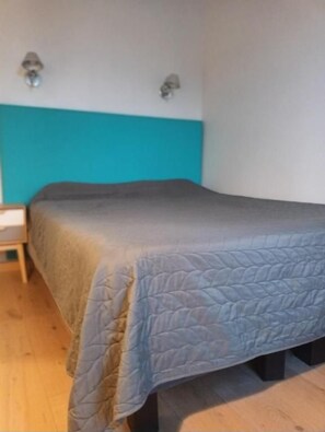 Una cama individual con cabecera apoyada en una pared, una mesita de noche a la izquierda, y una lámpara de pared sobre la cama.