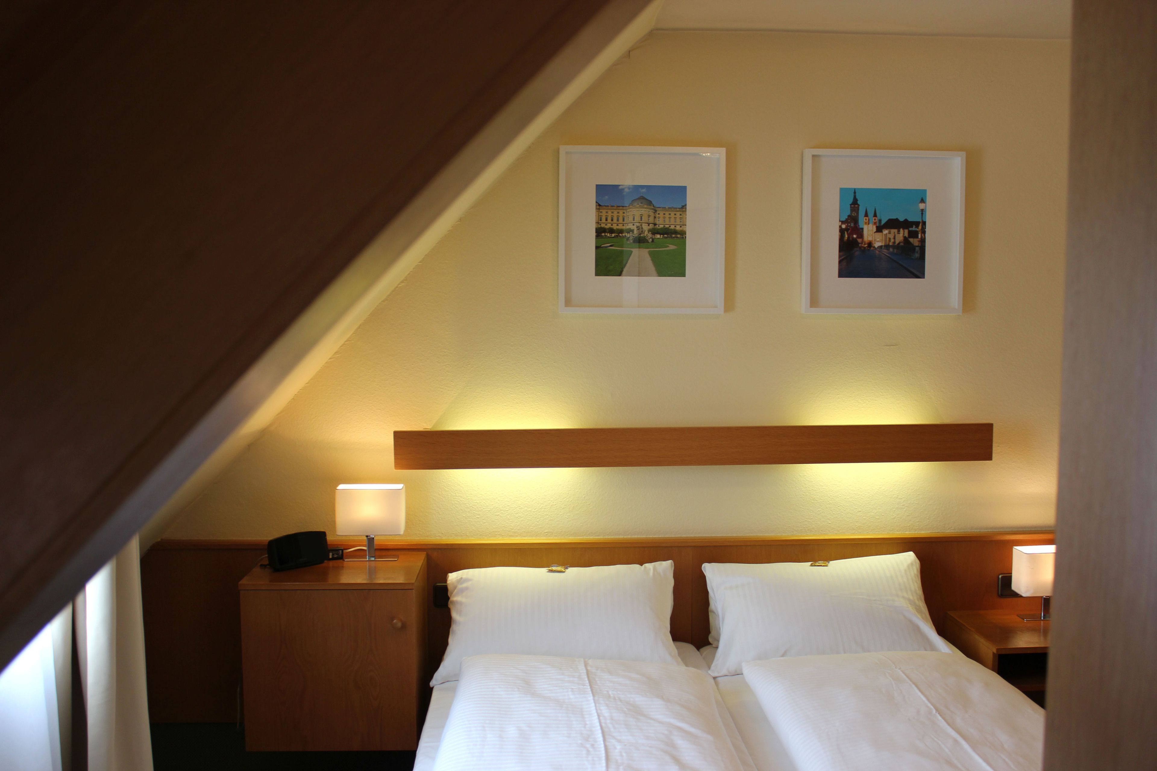 Camera d'albergo con una testiera in legno, due letti, un comodino con una lampada e due quadri appesi al muro.