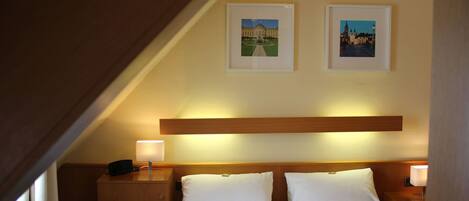 A hotel room with a wooden headboard, two beds, a nightstand with a lamp, and two framed pictures on the wall.