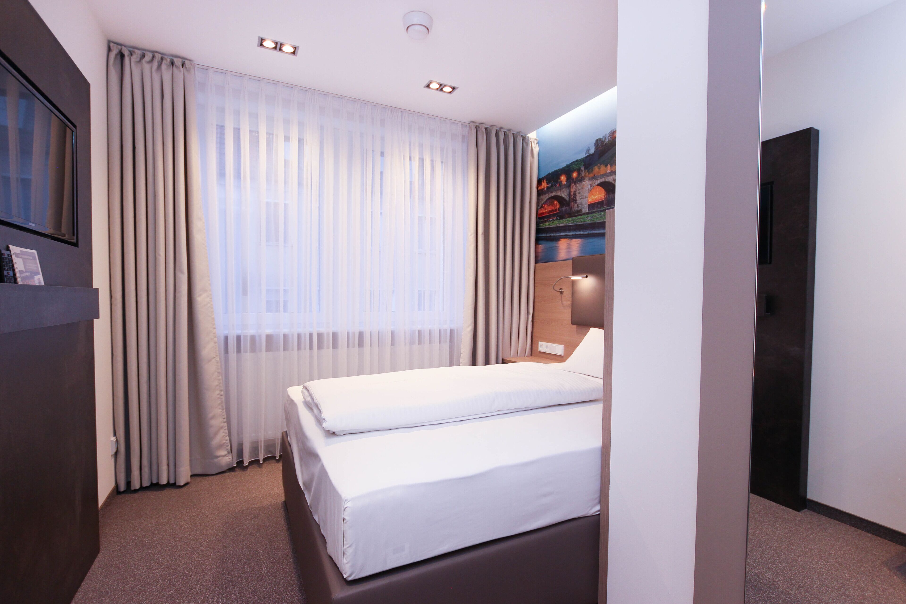 Ein Hotelzimmer mit einem Bett, Vorhängen, einem an der Wand befestigten Fernseher und einem Gemälde einer Brücke.