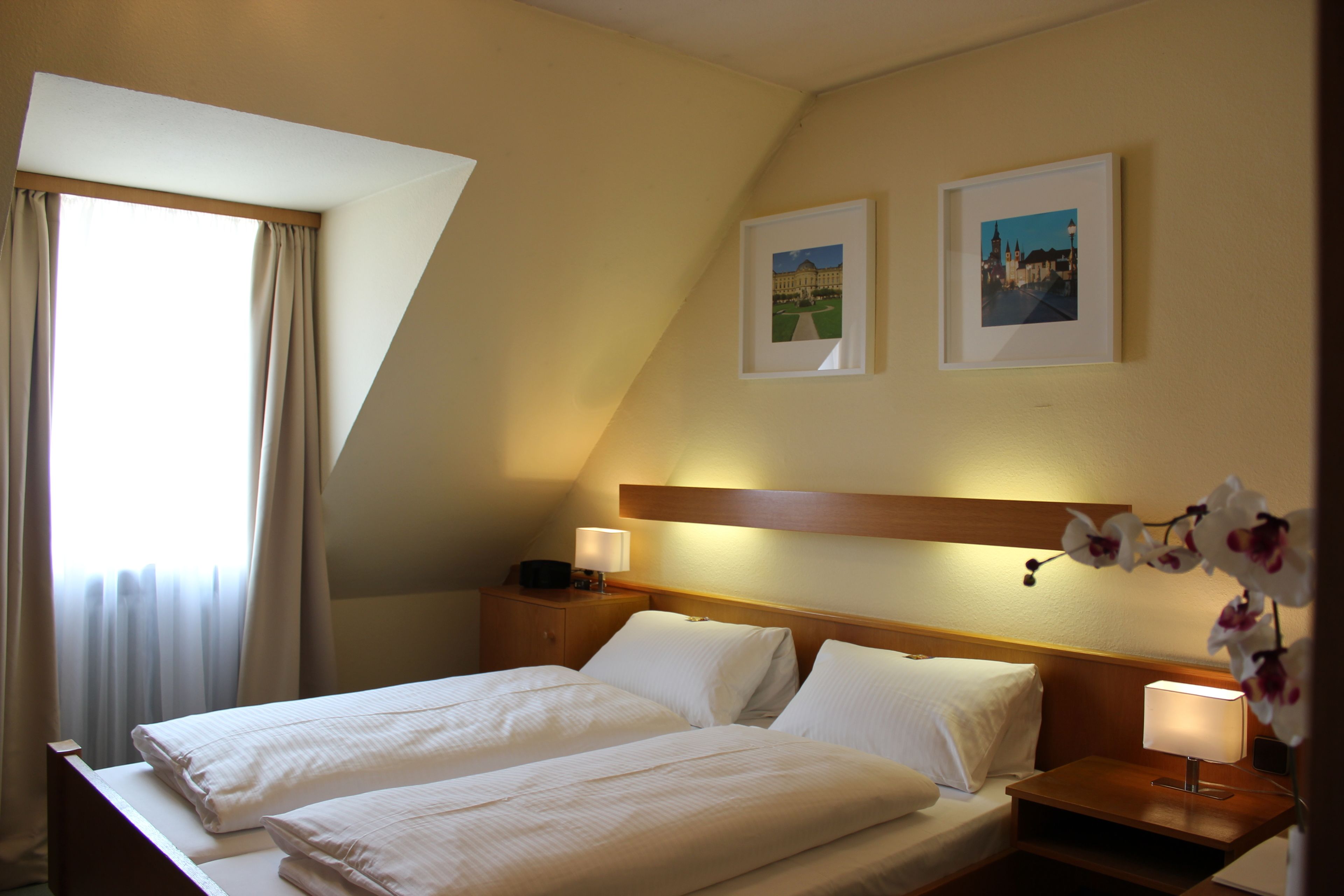 Habitación de hotel con dos camas, una ventana con cortinas, una mesita de noche con lámpara y dos cuadros enmarcados en la pared.