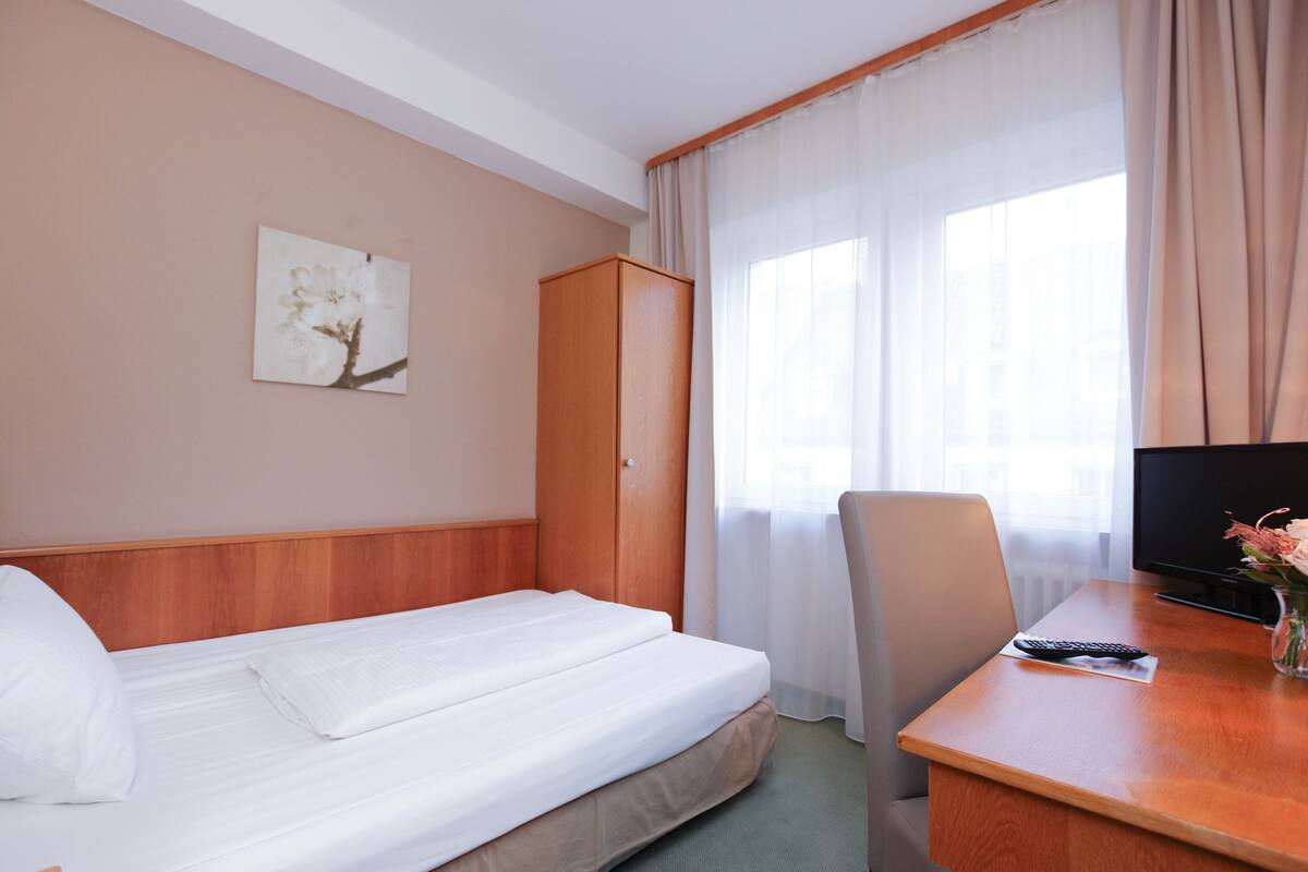 A hotel room with a bed, a desk with a remote control, a chair, a wardrobe, and a window with curtains.
