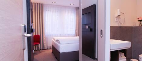 Habitación de hotel con cama, una silla roja, televisor, baño con lavamanos, inodoro y ducha.
