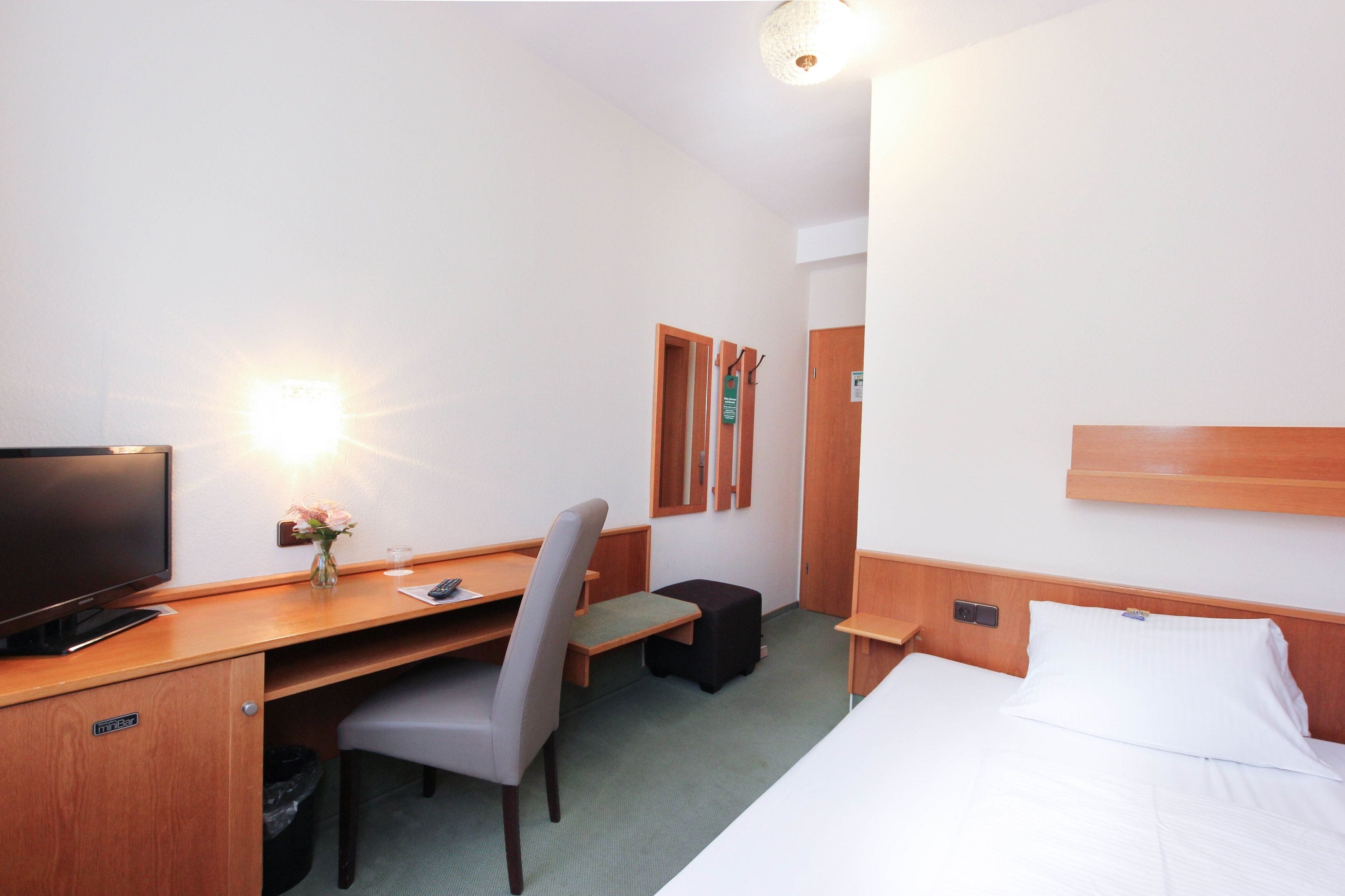 A hotel room with a bed, a desk with a computer, a chair, a TV, and a small table with a vase of flowers.