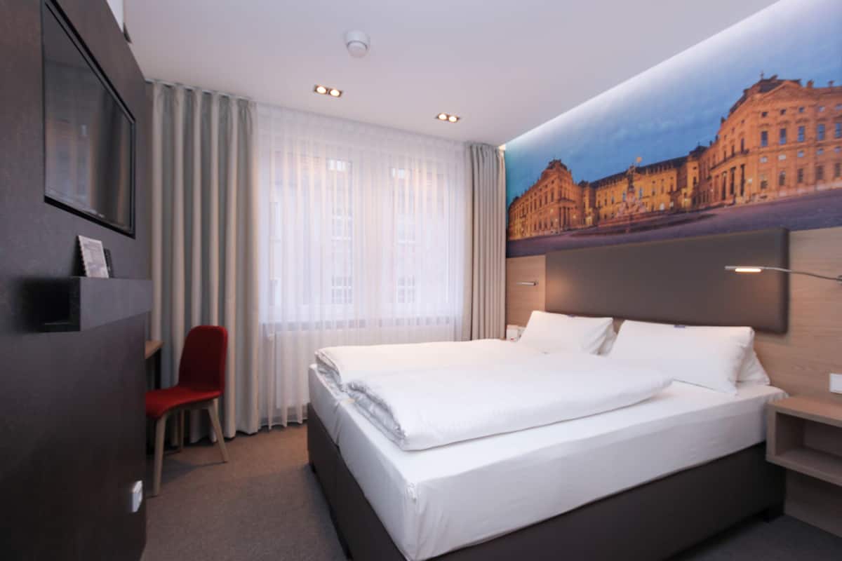 A hotel room with a large bed, a red chair, a TV, and a mural of a building on the wall.