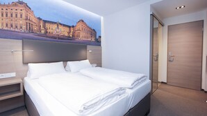 A modern hotel room with a large bed, a nightstand, a wooden headboard, and a view of a historic building through a large window. - City Hotel Würzburg (Würzburg)