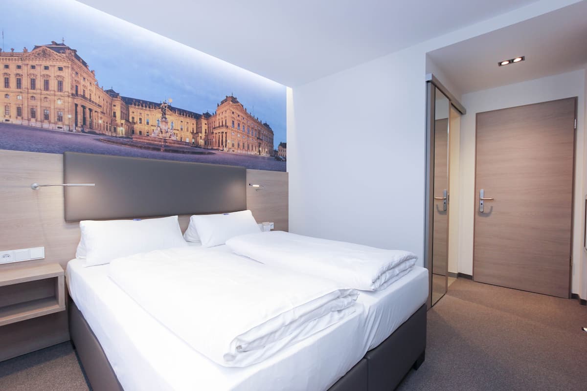 A modern hotel room with a large bed, a nightstand, a wooden headboard, and a view of a historic building through a large window.