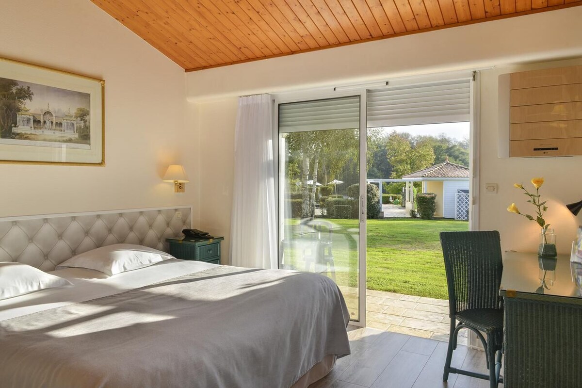 Un dormitorio con una cama grande, una mesita de noche, una lámpara y vistas a un jardín a través de un ventanal corredero.