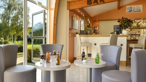 Zona de bar con vistas al exterior, incluyendo un campo de golf. Hay dos mesas redondas con sillas, y se ve la barra con varias botellas y una máquina de café.