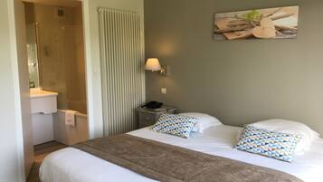 Habitación de hotel con una cama grande, una mesita de noche, una lámpara, un radiador, un cuadro en la pared y un baño con un lavabo y un espejo.