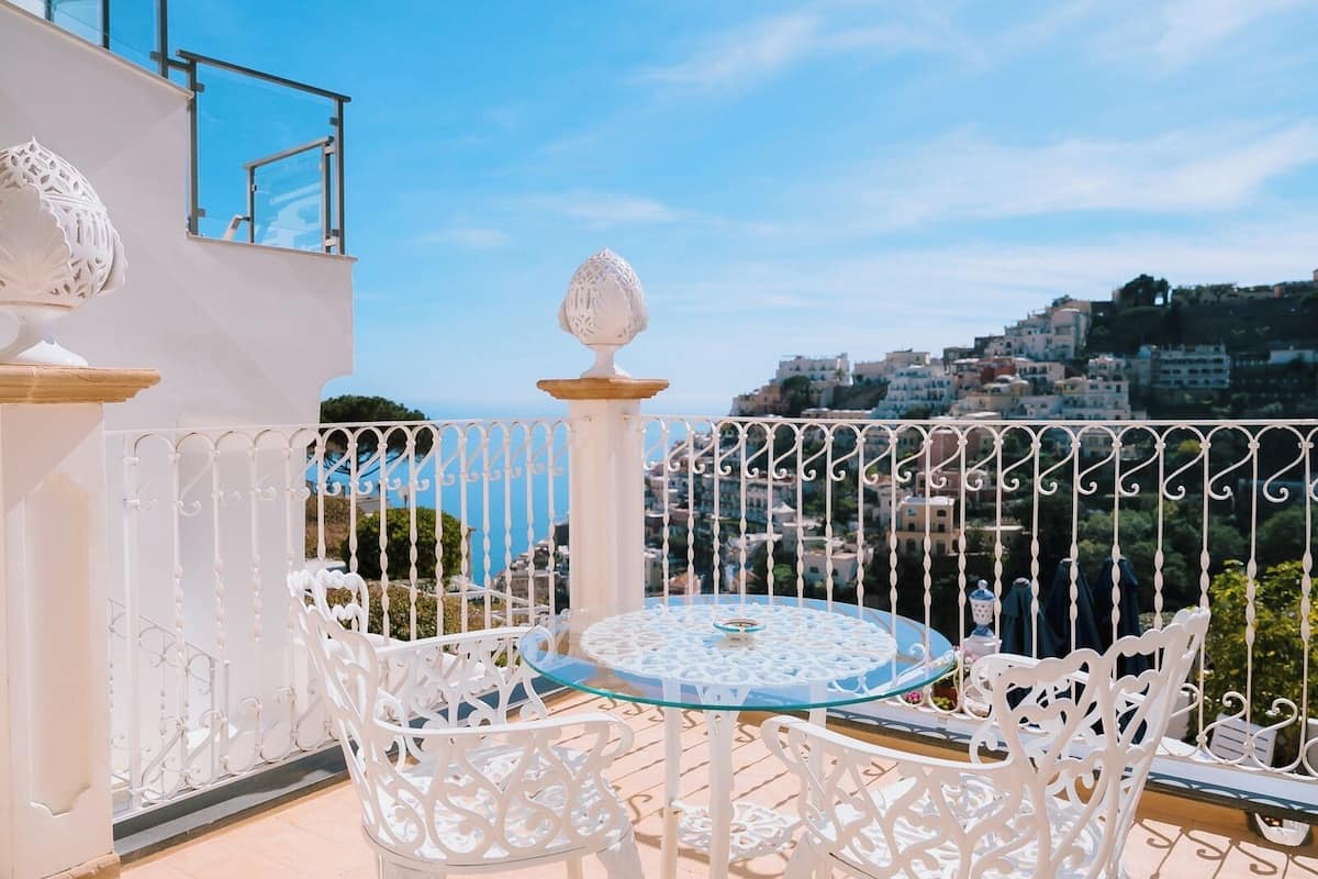Un balcón con una mesa de vidrio y sillas blancas ornamentadas, con vista a una ciudad costera.