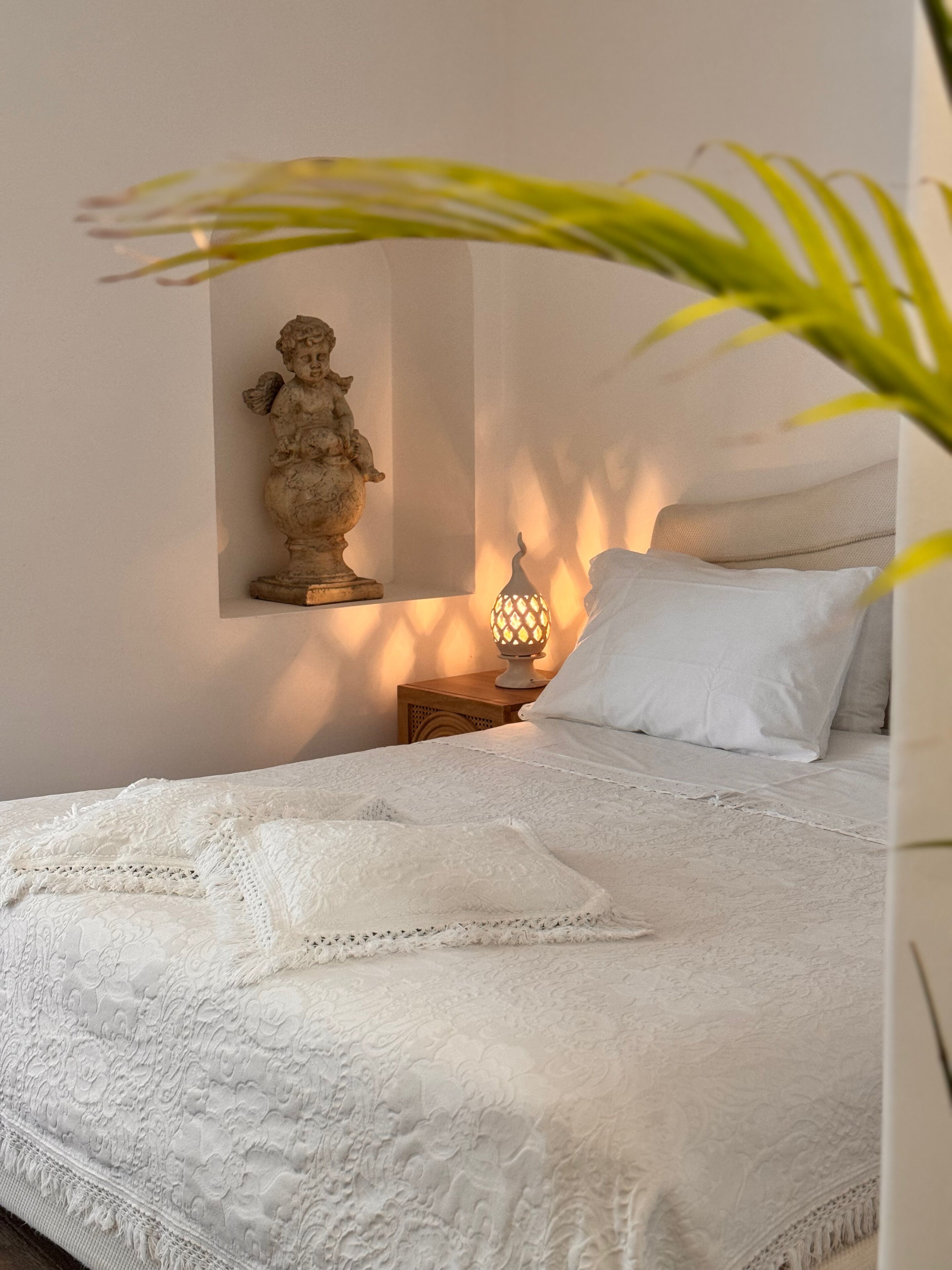 A neatly made bed with a white bedspread and pillows, a small wooden nightstand with a decorative lamp, and a wall-mounted sculpture.