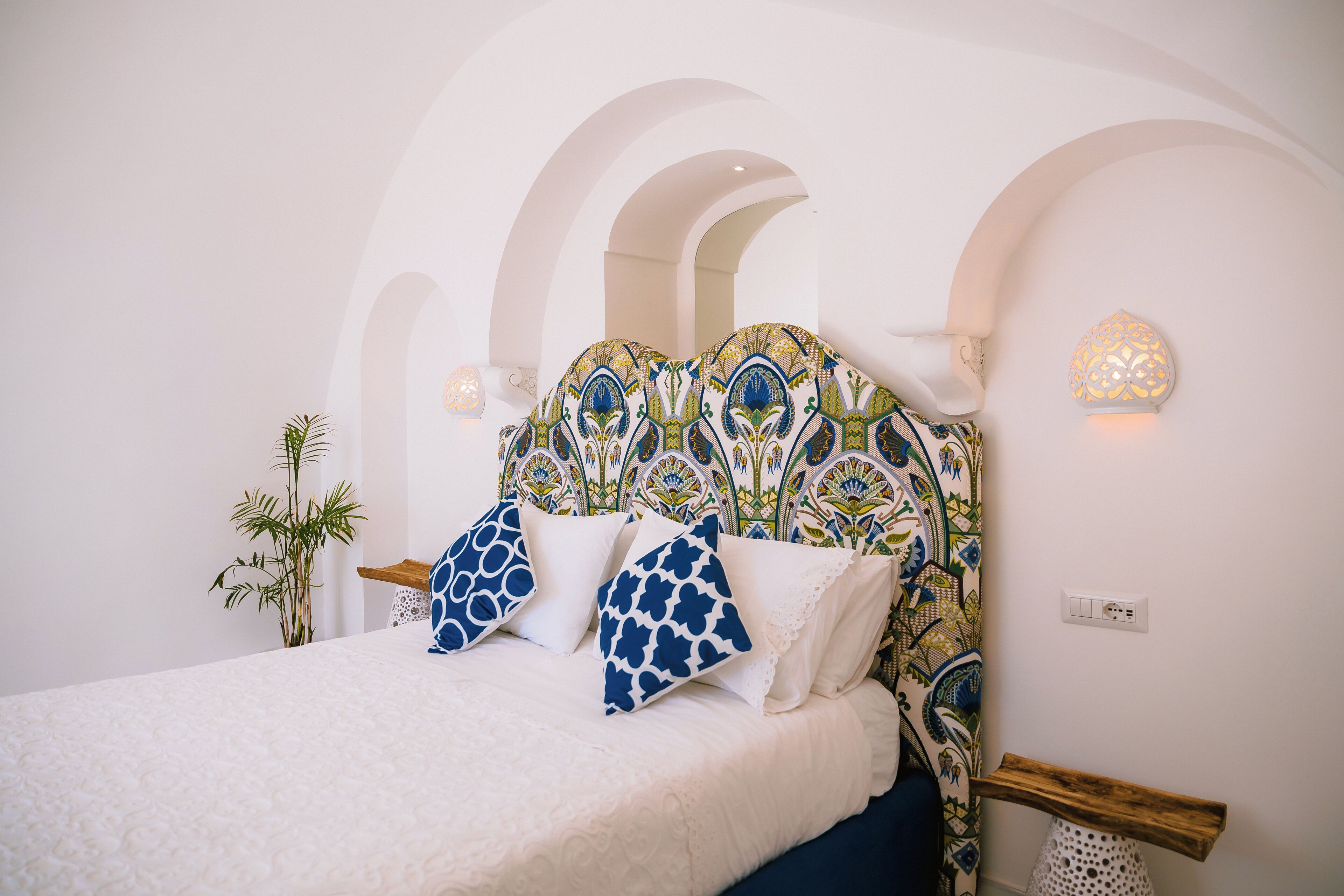 A bed with a patterned headboard, a potted plant, and a wall-mounted light fixture.