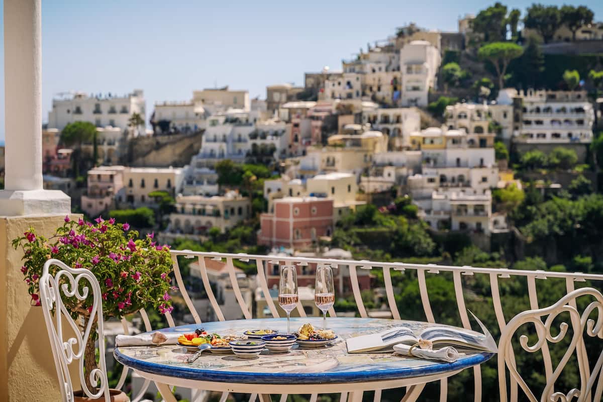 Una mesa con comida y bebidas en un balcón con vistas a una ladera de edificios muy cercanos.