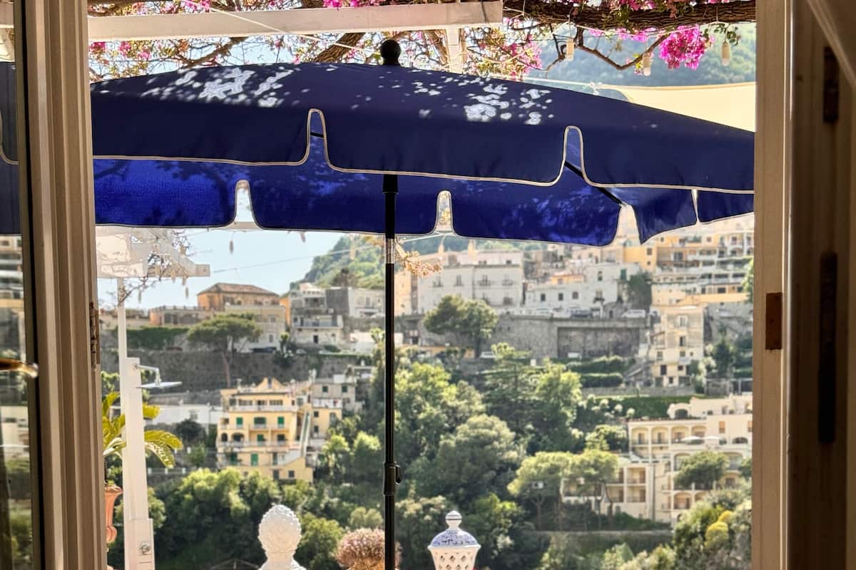 Un balcón con mesa y sillas, una sombrilla azul y vistas a un paisaje urbano con edificios y árboles.