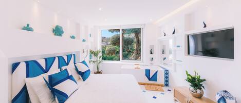 A modern bedroom with a large bed, a TV, and a window with a view of greenery.