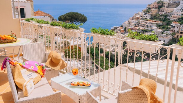 A balcony with wicker furniture, a table with a drink and food, and a view of a coastal town. - Villa Mary Suites (Positano)