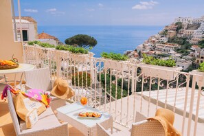 A balcony with wicker furniture, a table with a drink and food, and a view of a coastal town. - Villa Mary Suites (Positano)