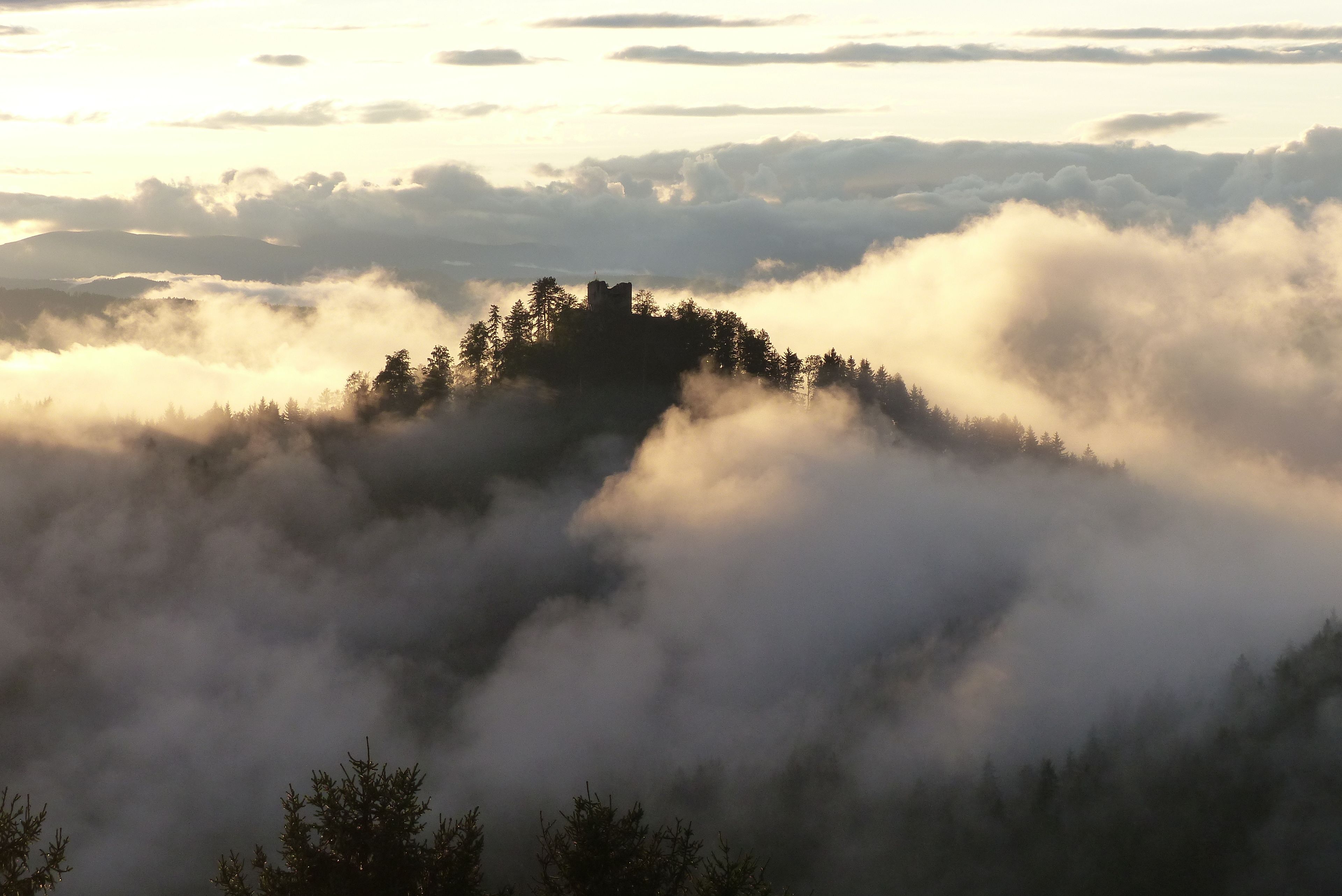 A forested hill with a castle-like structure surrounded by clouds.