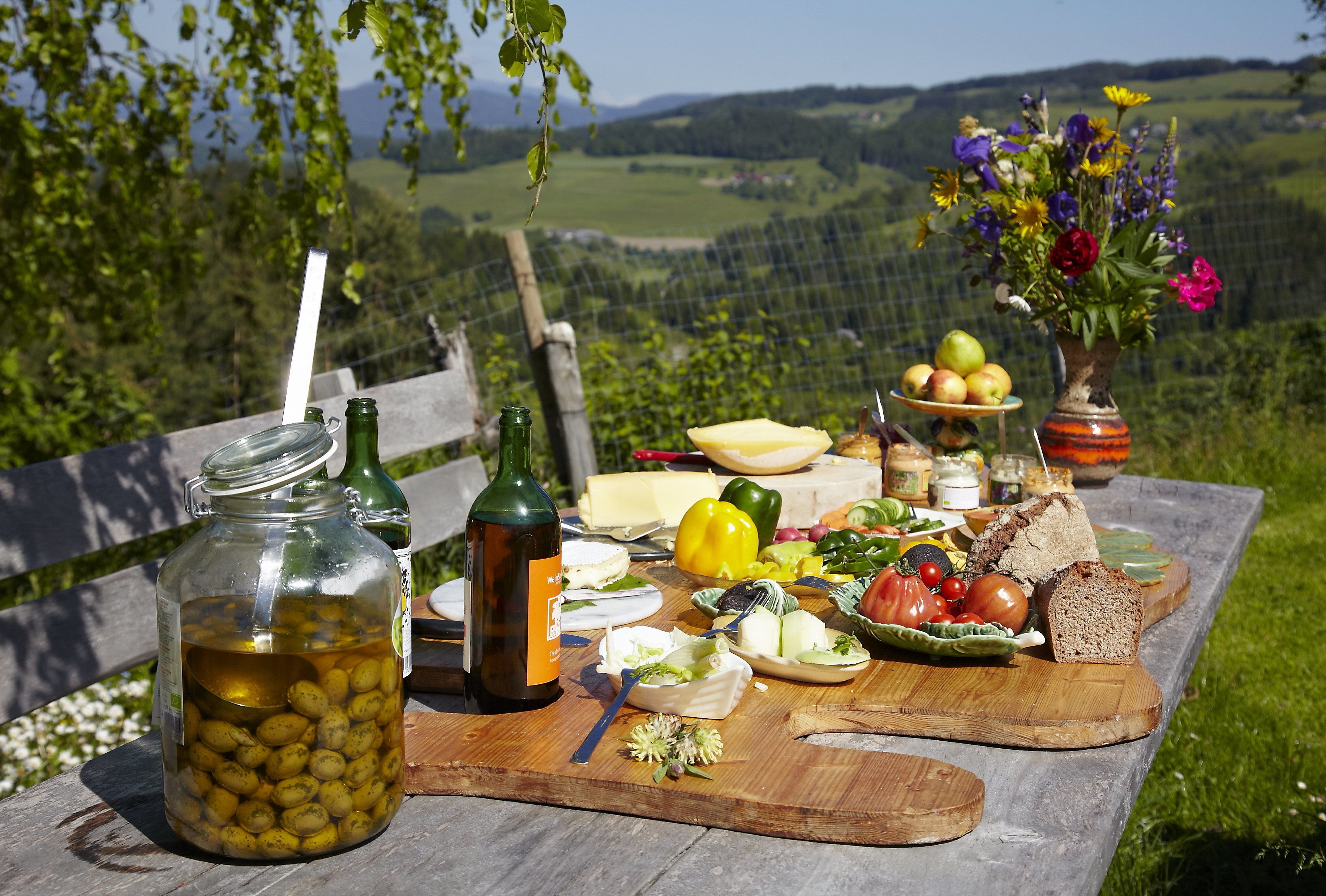 En picknickuppsättning med ett träbord, en burk med oliver, en flaska vin, en vas med blommor och diverse matvaror.