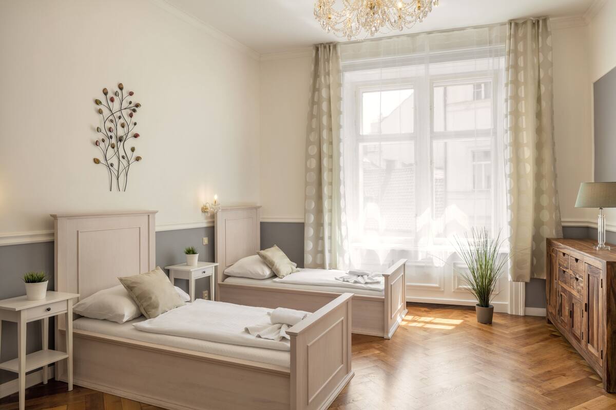 A bedroom with two beds, a chandelier, a window with curtains, and a wooden cabinet.