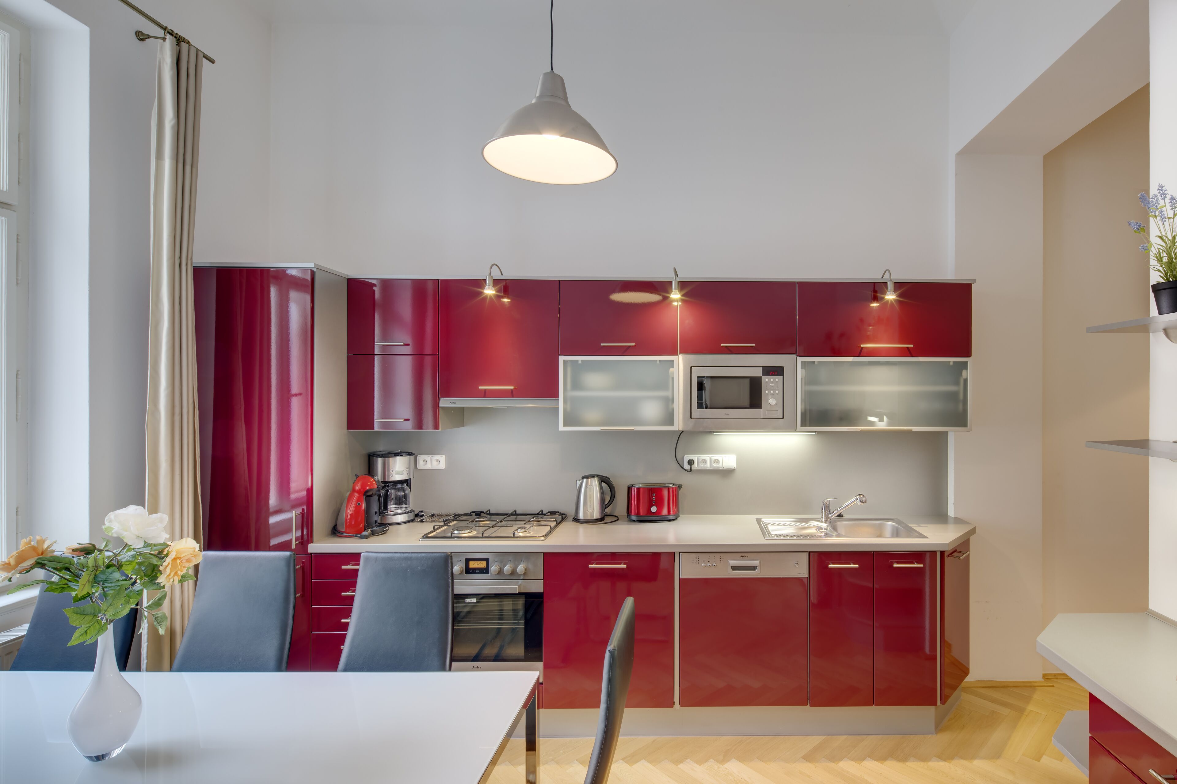 A modern kitchen with red cabinets, a white countertop, and a dining area with a table and chairs.