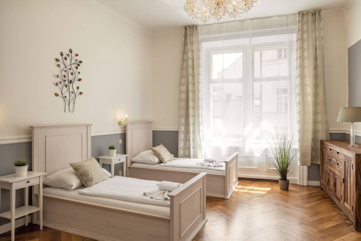 A bedroom with two beds, a chandelier, a window with curtains, a bedside table with a lamp, and a cabinet.