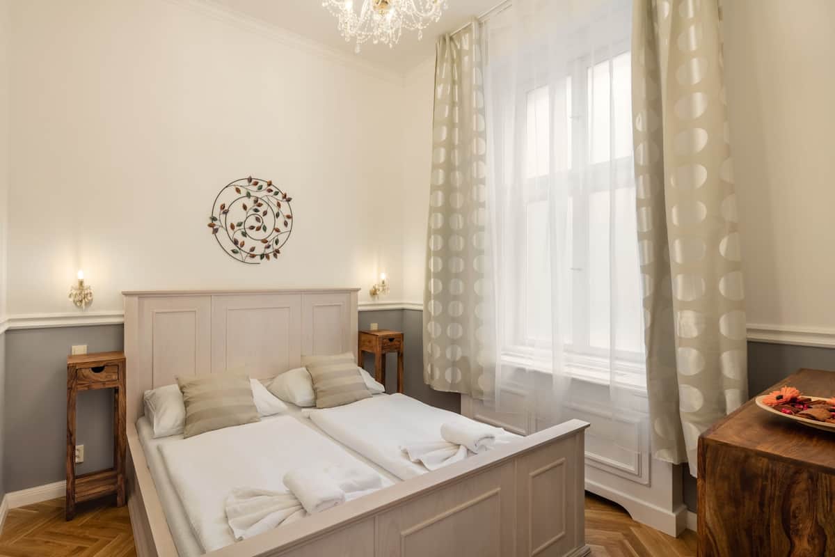 A double bed with a wooden headboard, two bedside tables, a chandelier, a decorative wall piece, a window with sheer curtains, and a wooden side table with a plate.