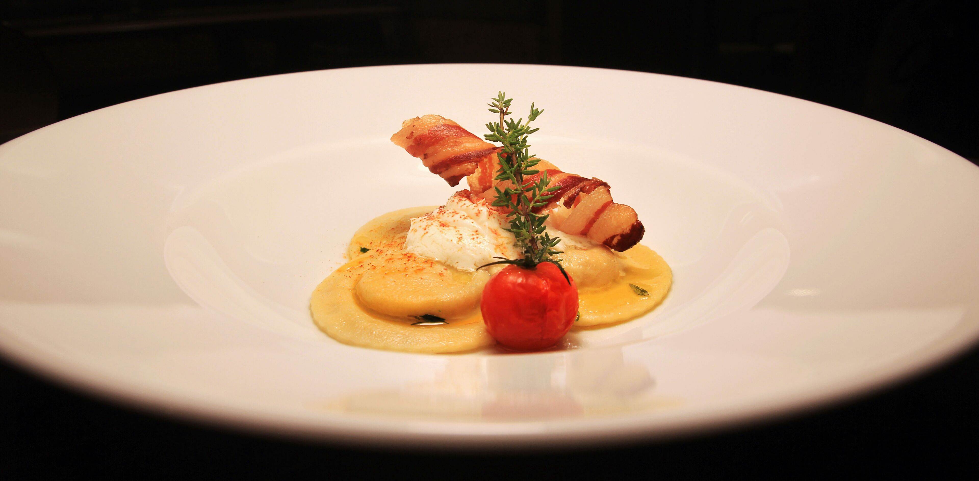 A white plate with a dish consisting of a poached egg, a slice of bacon, a tomato, and a garnish of thyme.