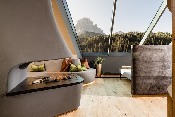 A modern living room with a curved sofa, a small table, and a view of a mountainous landscape through large windows. - Hotel Touring (Santa Cristina Val Gardena)