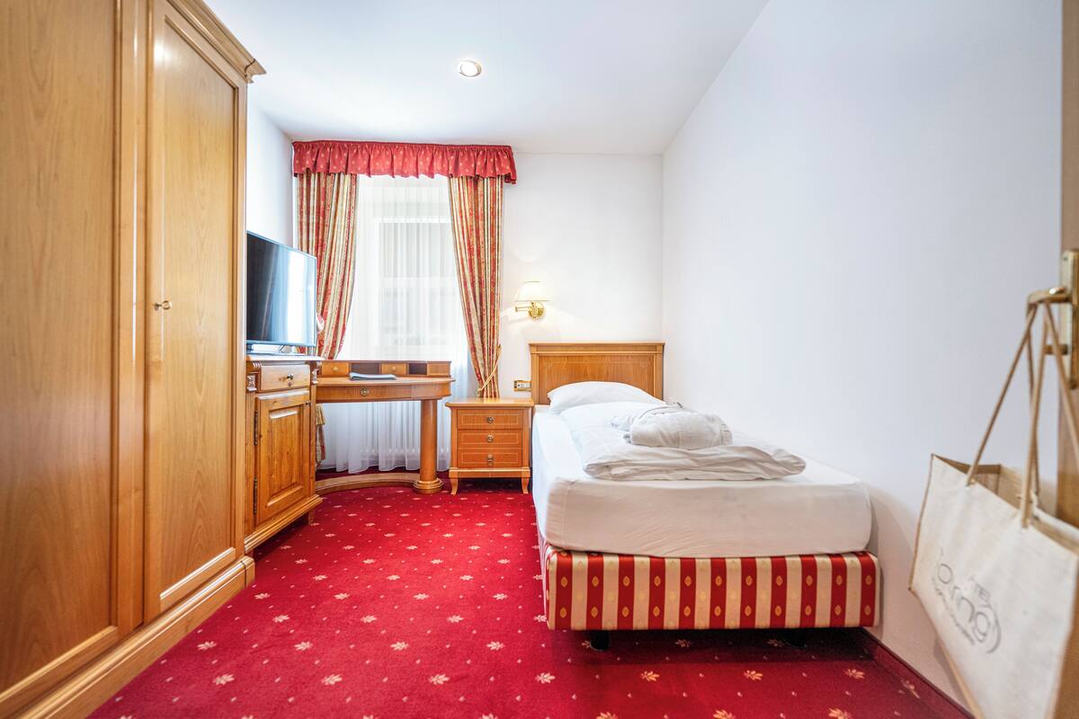 Une chambre d’hôtel avec une armoire en bois, un bureau avec une télévision, un lit avec une tête de lit à rayures et une moquette rouge.