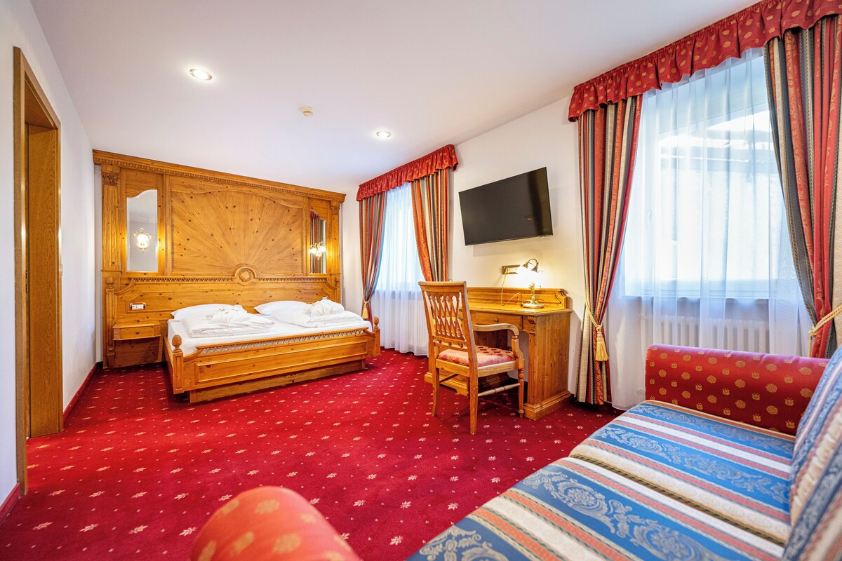Une chambre d’hôtel avec un lit en bois, un bureau, une chaise, une télévision et une grande fenêtre avec des rideaux.