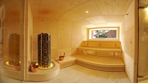 Una sauna de madera con un pilar central de piedra, bancos y una ventana que da a un muro de piedra.
