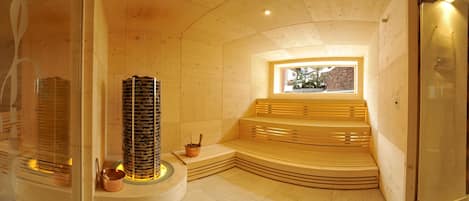 Una sauna de madera con un pilar central de piedra, bancos y una ventana que da a un muro de piedra.
