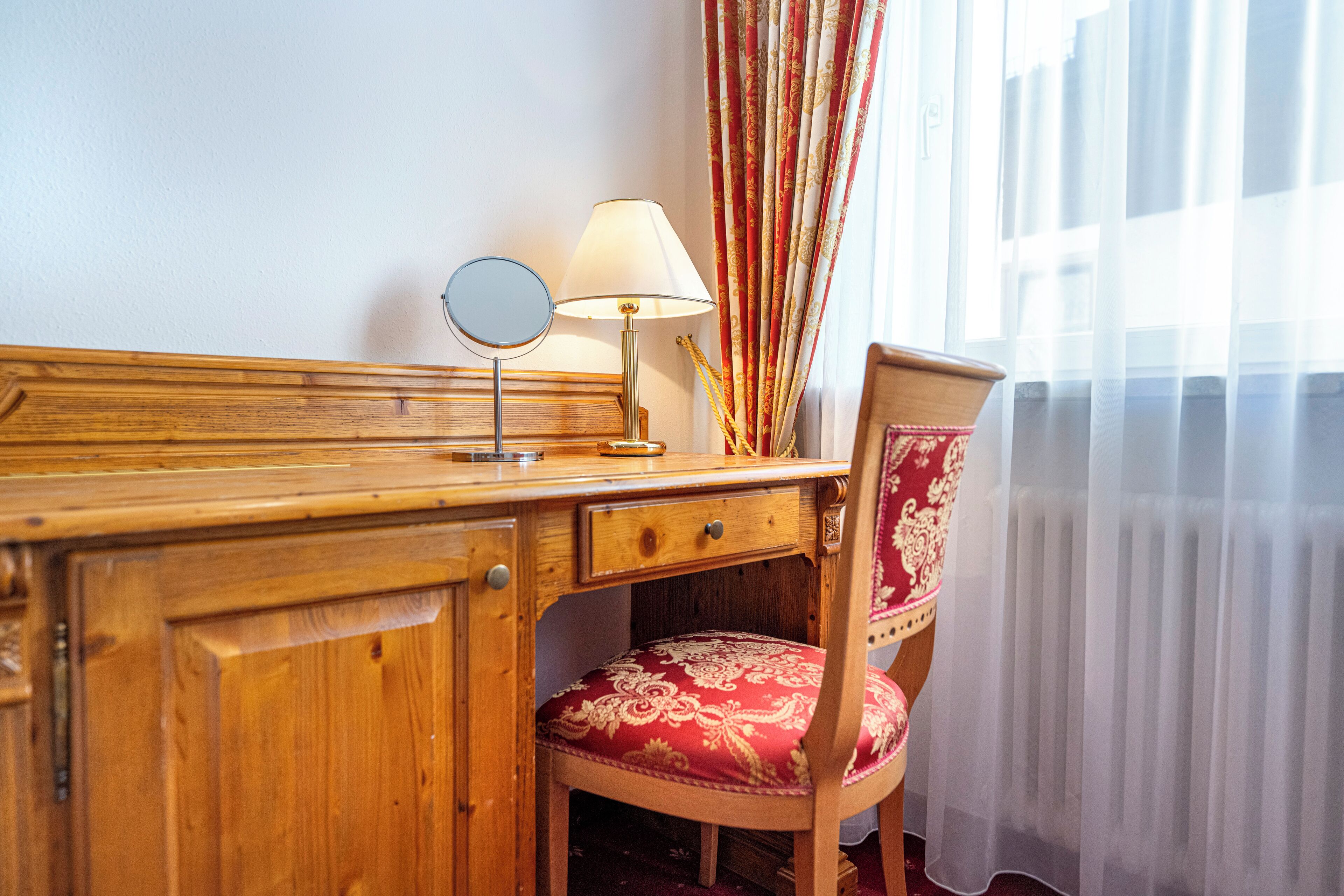 Een houten bureau met een lamp, een spiegel en een rode fauteuil.
