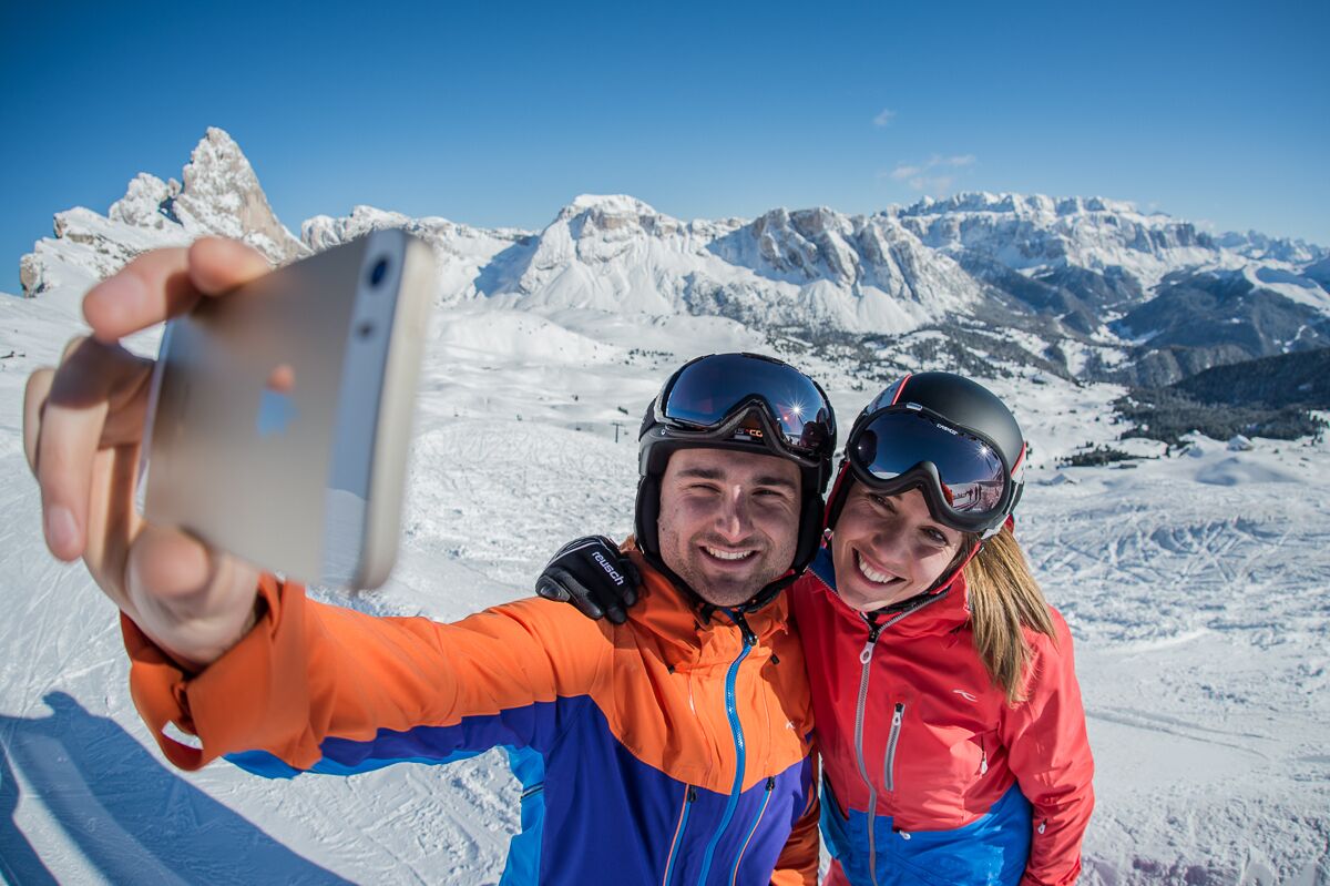 A person taking a selfie with a smartphone, wearing a red and blue jacket, and a helmet, with a snowy mountain landscape in the background.