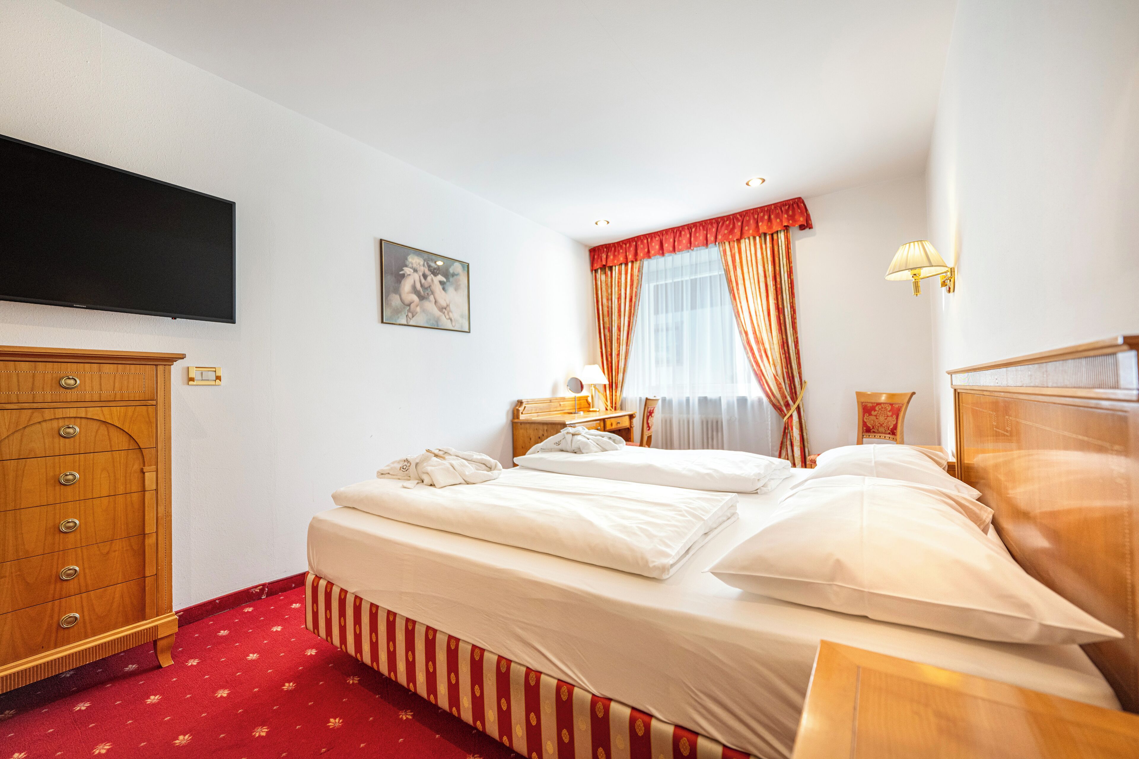 A hotel room with a bed, wooden wardrobe, flat-screen TV, artwork, and a window with curtains.