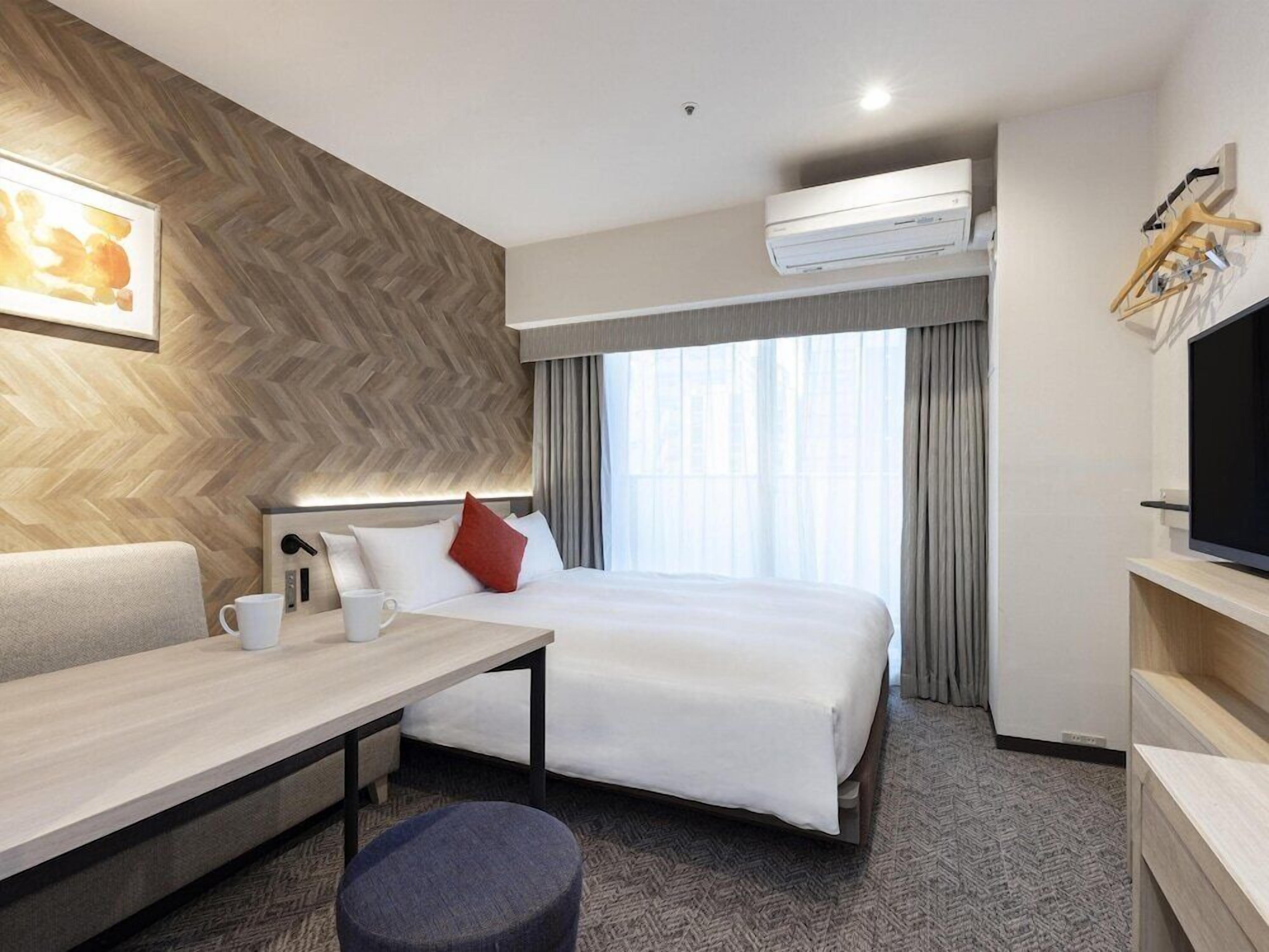 A hotel room with a bed, a desk, a chair, a television, and an air conditioning unit.